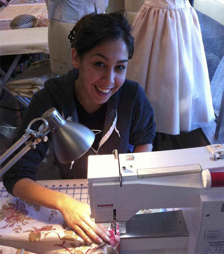 A female student with dark hair sews at a sewing machine and smiles at the camera