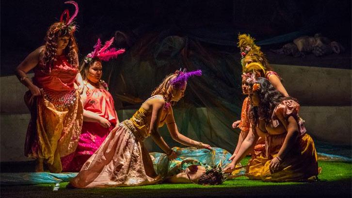 Actors in bright yellow, pink and purple fairy costumes surround an actor on a grassy stage in a Ventura College production of A Midsummer Night's Dream.